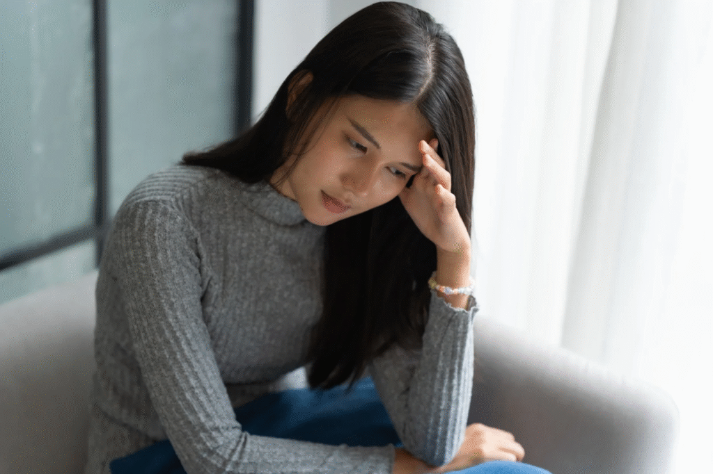 A picture showing a lady sitting on a couch by the window, processing her thoughts after finding out her partner has a lot of debts