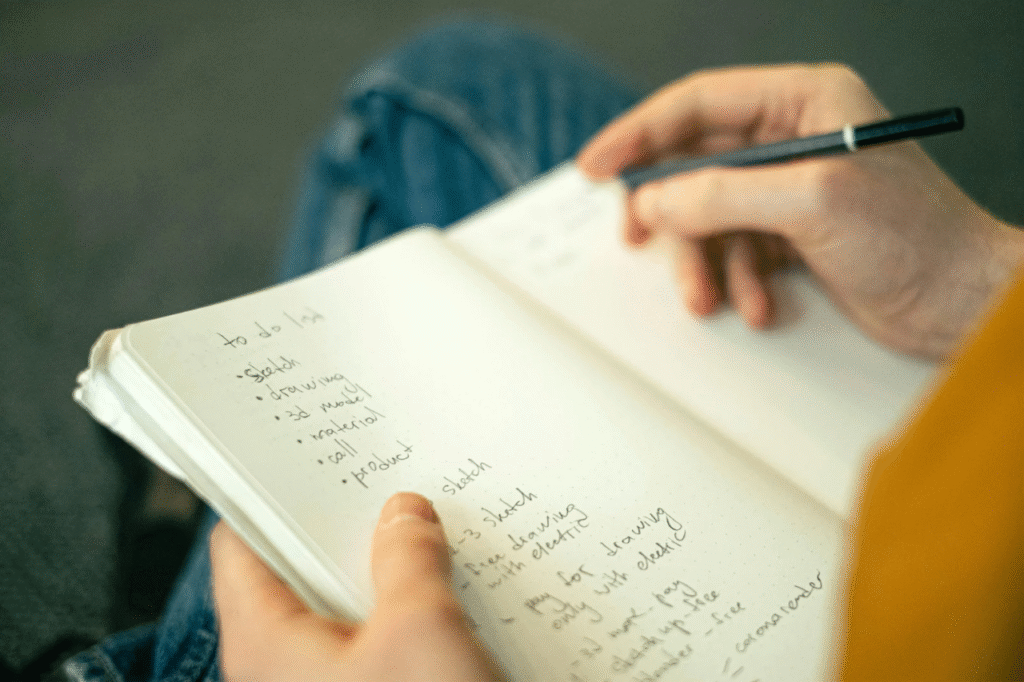 A picture Close-up of hands writing in a notebook with a laptop nearby, listing different debts, balances, and interest rates.