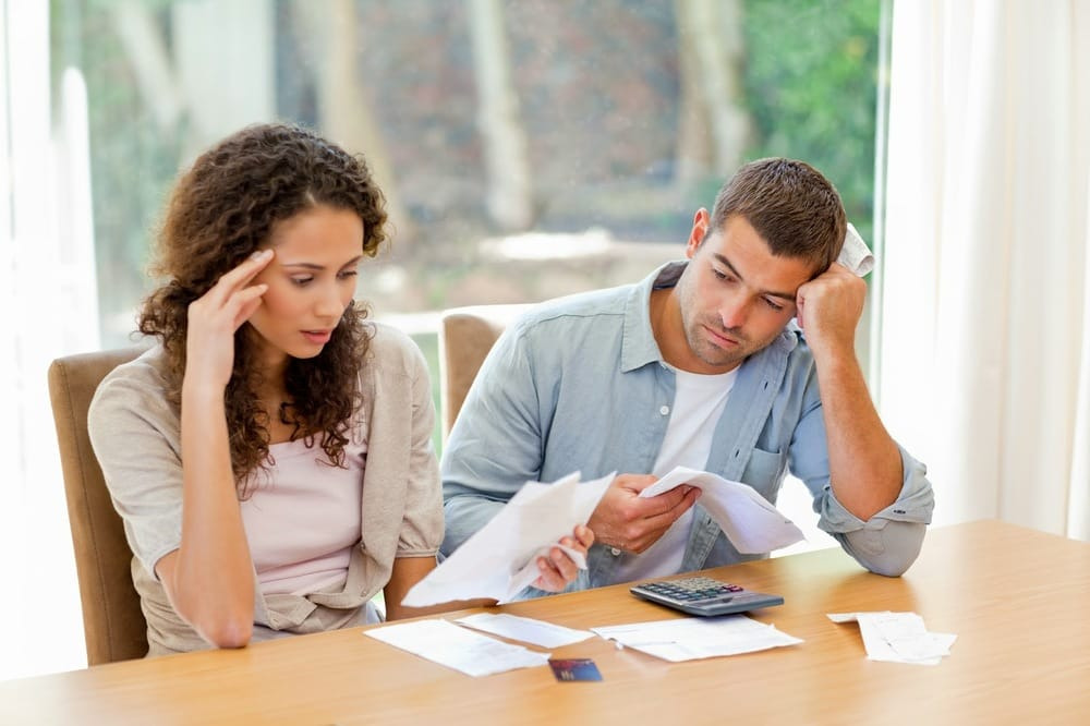 A picture showing couple sitting together at a table, looking concerned while reviewing financial documents, representing the challenges of managing money when a partner has a lot of debt