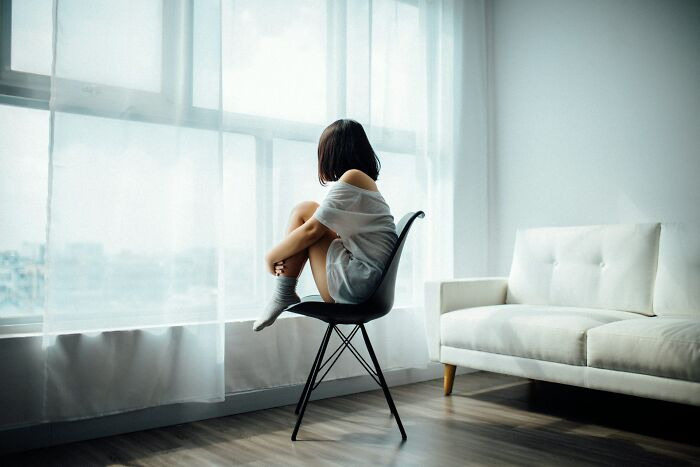 An image showing a woman sitting alone on a chair facing a window in a sparsely furnished room, illustrating the emotional withdrawal and silence that grows between couples due to financial mistakes 