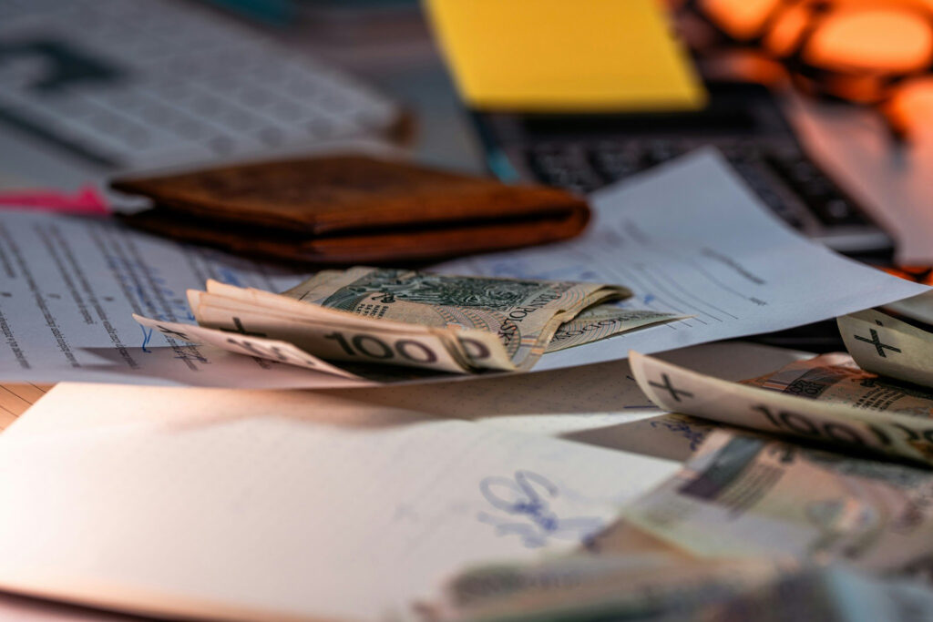 An image showing A  cluttered desk with a brown wallet, stacks of banknotes, and various documents under warm lighting.